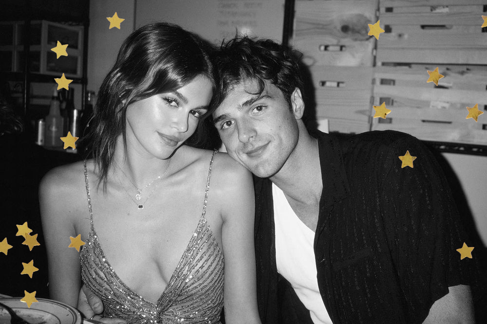 Foto em preto e branco de Kaia Gerber e Jacob Elordi; ele está encostando a cabeça no ombro de Kaia e os dois sorriem levemente; ela usa um vestido de alças e ele usa camiseta branca com uma camisa escura por cima; estrelas amarelas decoram a imagem