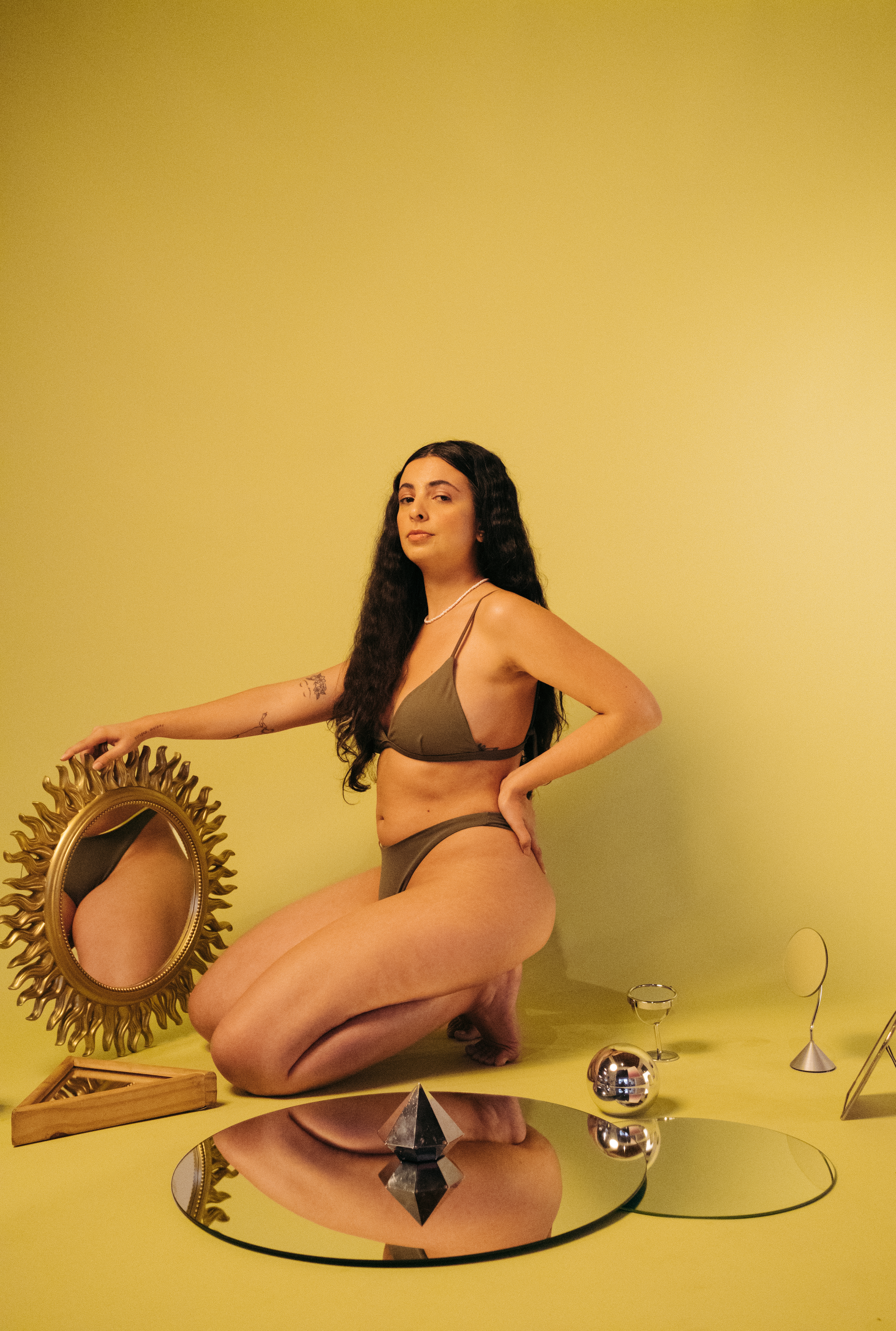Ilustradora Lela Brandão usando biquíni marrom da sua própria marca, agachada com uma das mãos no quadril, segurando um espelho e com expressão facial séria