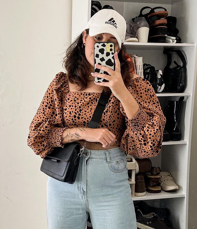 Selfie de uma mulher no espelho. Ela usa uma calça mom jeans, blusa com manga bufante com estampa de animal, boné com aba curva branco e bolsa preta. Ela olha para o celular e não sorri.