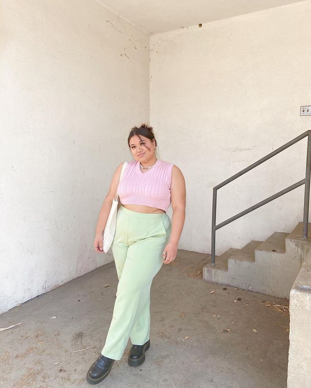 Foto de uma mulher próxima a uma escada. Ela usa um colete de vovô rosa, calça verde, coturno preto e ecobag. Ela está com o cabelo preso em um coque baixo, olha para o lado e sorri levemente.