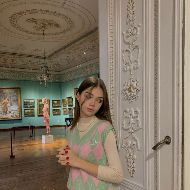 Foto de uma menina em um museu. Ela usa um suéter bege, colete de vovó em tons de verde e rosa como sobreposição e colar de pérolas. Ela olha para o lado e não sorri para a foto.