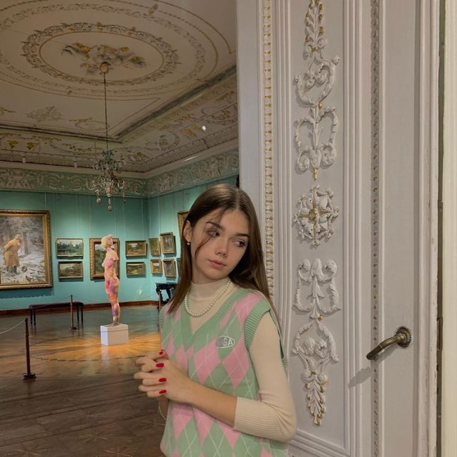 look-colete-de-vovo Foto de uma menina em um museu. Ela usa um suéter bege, colete de vovó em tons de verde e rosa como sobreposição e colar de pérolas. Ela olha para o lado e não sorri para a foto.