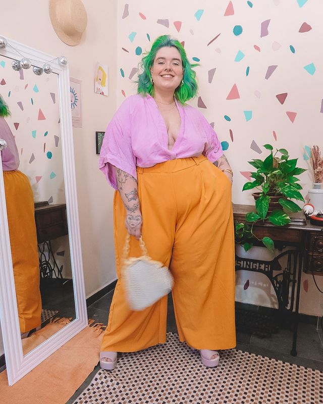 Foto de uma mulher em um quarto. Ela usa uma camisa lilás, calça laranja, tamanco rosa e bolsa branca. Ela está com o cabelo curto e verde solto, olha para a câmera e sorri.