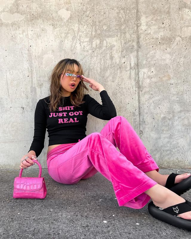 Foto de uma mulher sentada no chão. Ela usa uma blusa de manga longa preta com detalhes rosa, calça rosa, chinelo preto, bolsa rosa e óculos de sol. Ela está com a mão esquerda no óculos e segura a bolsa com a mão direita, olha para o lado e mostra a língua.