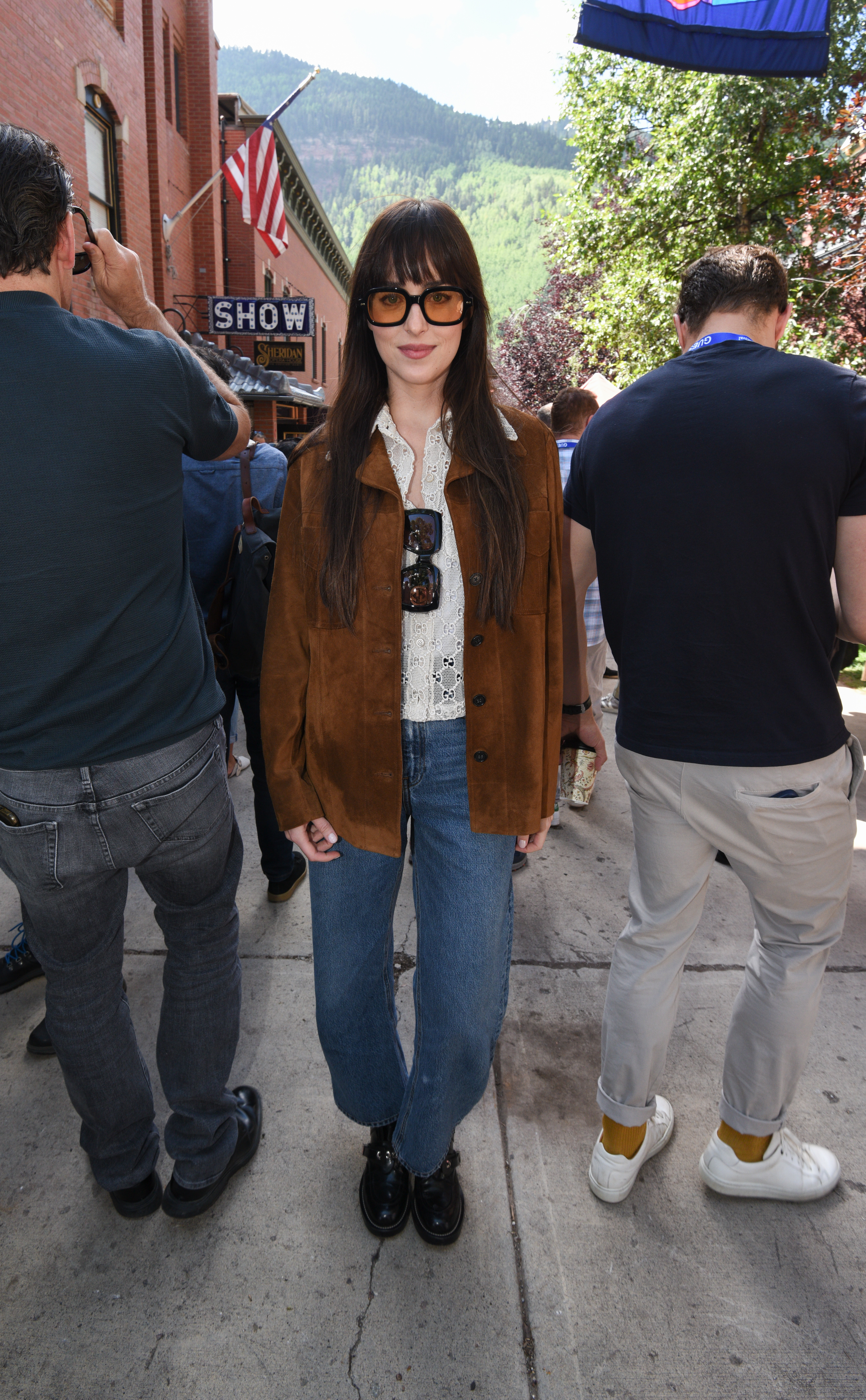 Dakota Johnson usando óculos de lentes amareladas, camisa branca, jaqueta marrom de camurça, calça jeans e bota preta na rua, ao lado de pessoas viradas de costas