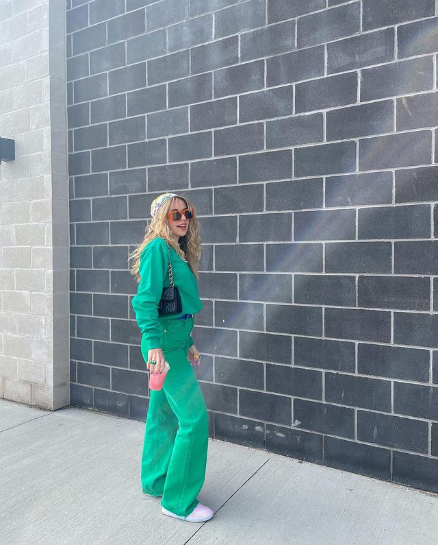 Foto de uma mulher em uma calçada. Ela usa um moletom verde, calça verde, tênis branco, lenço colorido no cabelo, bolsa baguete preta e óculos de sol com armação vermelha. Ela segura um copo na mão direita, olha para a frente e sorri levemente.