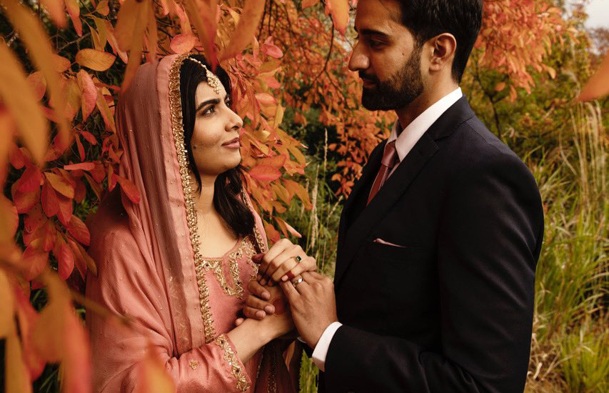 Foto do casamento de Malala. A paquistanesa veste uma roupa coral, está olhando para o marido e segura as mãos dele
