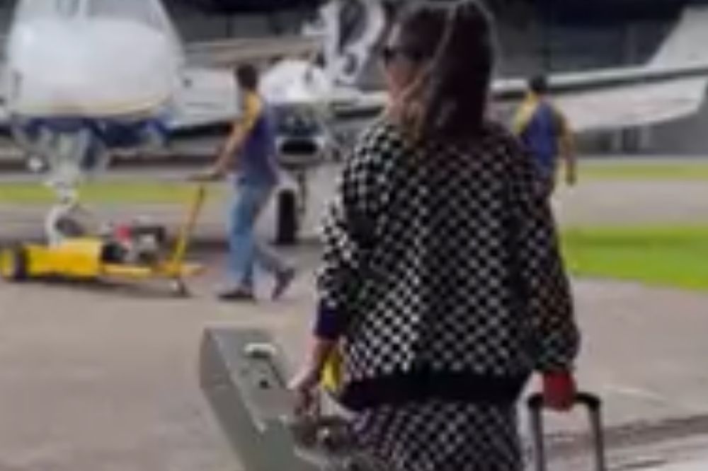 Marília Mendonça de costas segurando uma mala e uma capa de violão; ao fundo é possível ver uma aeronave branca com detalhes em azul e amarelo; ela usa um casaco quadriculado nas cores branco e preto e óculos escuro preso com cabelo preso