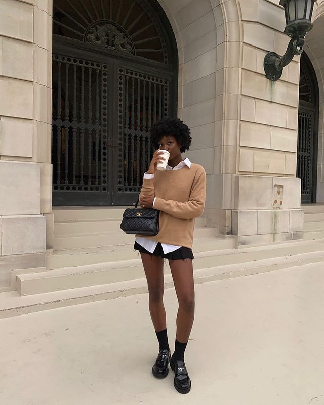 moda-preppy Foto de uma mulher em uma calçada. Ela usa uma camisa branca, colete de vovó bege como sobreposição, saia preta, mocassim preto e bolsa preta. Ela segura um copo de café na mão direita, olha para a câmera e não sorri.