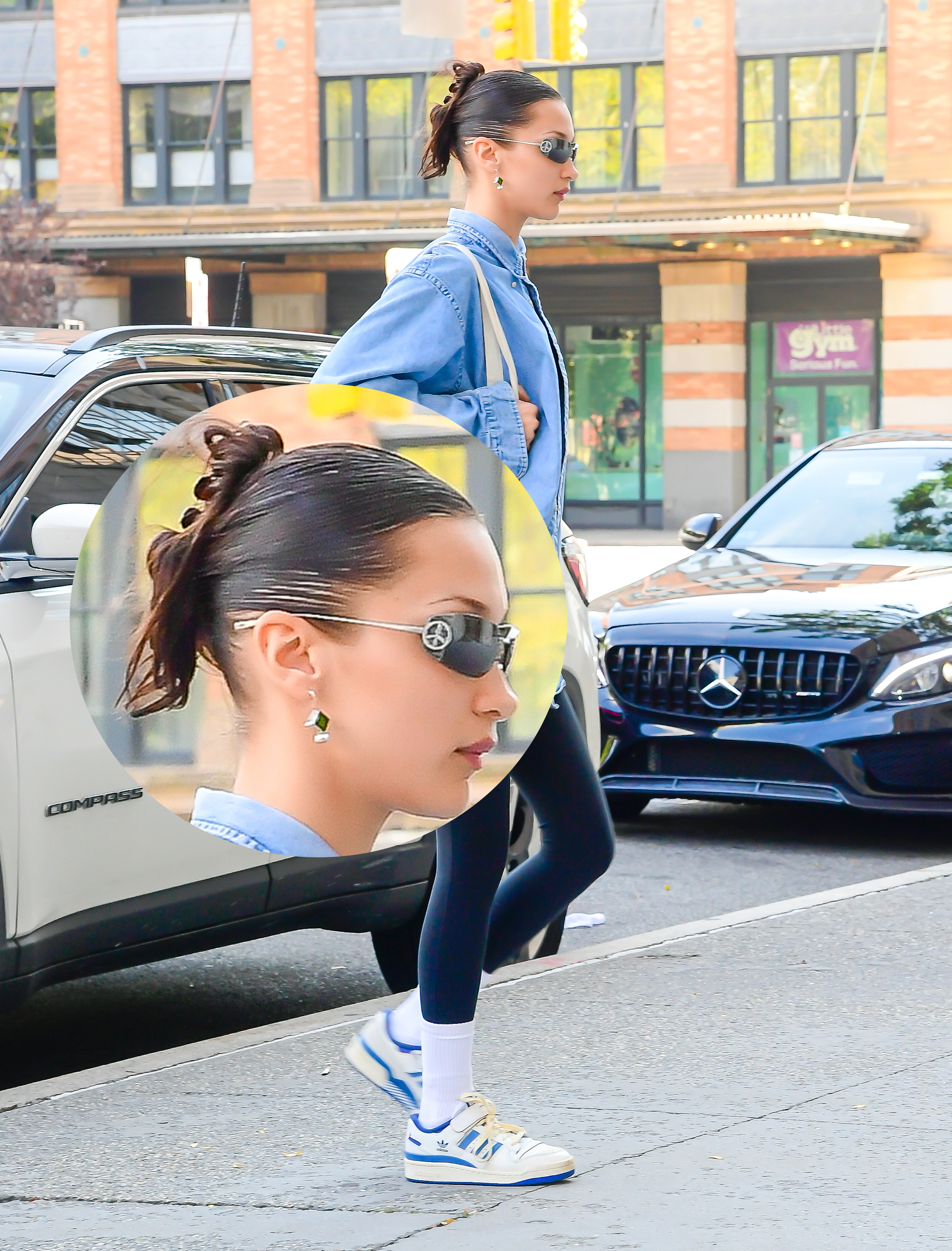 Bella Hadid andando na rua com camisa azul, legging azul-marinho, tênis branco, óculos de sol e penteado com piranha de cabelo