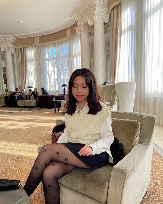 preppy Foto de uma mulher sentada em uma poltrona em uma sala. Ela usa uma camisa branca com colete de vovó como sobreposição bege, saia preta e meia calça preta. Ela olha para baixo e não sorri.