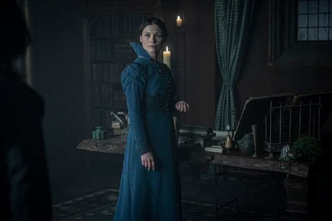 WW_102_09.11.2018_396.NEF Tissaia, interpretada por MyAnna Buring, em The Witcher, da Netflix; ela está em pé usando um vestido verde azulado; a personagem está parada no meio de um cômodo olhando de lado com uma das mãos levantadas próxima ao corpo e sua expressão é séria; as paredes são de madeira, a cortina é verde e velas iluminam o local que tem uma mesa próxima da atriz