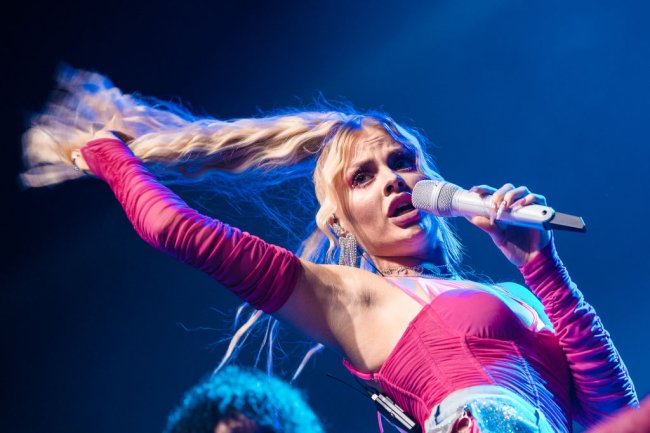 Foto de Luísa Sonza durante show. Ela está no palco, usa uma blusa sem alças e luvas pink, e segura o rabo de cabalo loiro com a ponta dos dedos