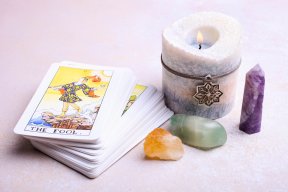 Tarot e amor: o que significa quando estas cartas saem no seu jogo? Baralho de Tarot organizado sobre uma mesa branca. A carta virada para cima é a do Louco. Ao redor do deck, uma vela acesa e pedras mineiras laranja e verde