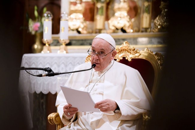 Foto do Papa Francisco sentado em uma cadeira chique, dentro de uma igreja, falando no microfone