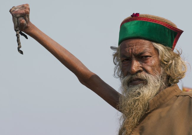 Foto do monge indiano Amar Bharati, que há mais de 45 anos vive com um braço esticado para o alto. O membro direito dele está atrofiado, as unhas estão gigantes, ele tem uma barba longa branca e veste um chapéu cocô vermelho e verde