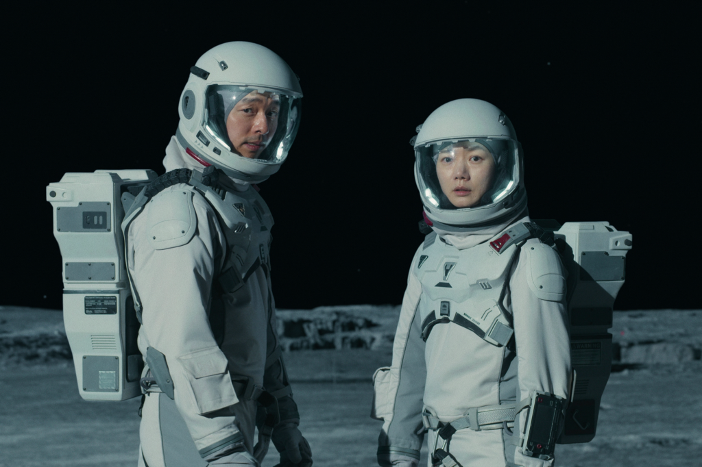 Imagem de divulgação de O Mar da Traquilidade, série da Netflix, com duas pessoas, um homem e uma mulher, usando trajes de astronauta brancos com capacete de material transparente na parte da frente do rosto; eles estão com expressões assustadas/confusas e ao fundo o céu está completamente escuro