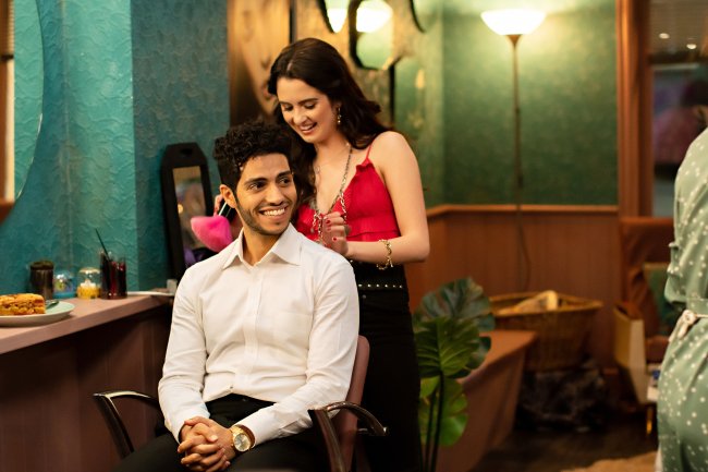 Laura Marano como Izzy e Mena Massoud como princípe Thomas em Tratamento de Realeza, da Netflix; ele está sentando sorrindo e olhando levemente para o lado enquanto ela está em pé atrás dele mexendo em seu cabelo para arrumá-lo