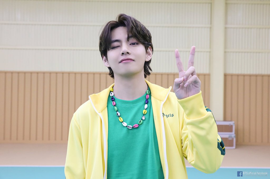 V do BTS com uma camiseta verde e um casaco amarelo com dois dedos levantados [indicador e anelar] de uma das mãos e piscando para a câmera enquanto posa para foto de divulgação