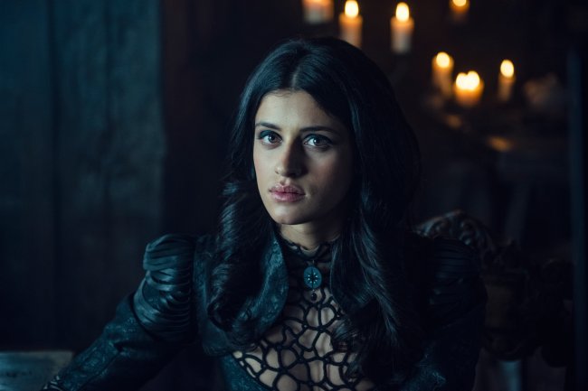 WW_105_20.02.2019_68.NEF Yennefer de Venberg, interpretada por Anya Chalotra, em The Witcher, da Netflix; ela está olhando para câmera com expressão neutra e usa um vestido preto com o cabelo solto dividido ao meio; ela está em um ambiente clareado pela luz de uma janela com um corredor escuro ao fundo com velas como iluminação