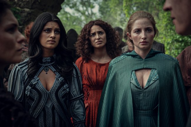 Imagem de Calanthe, Triss e Fringilla interpretadas por Jodhi May, Anna Shaffer e Mimi M Khayisa em The Witcher, da Netflix; as três estão com expressões confusas olhando para algo; a primeira da esquerda para direta veste um vestido azul claro com detalhes em azul escuro, a do meio um vestido alaranjado e a última um vestido verde; elas estão em paradas em uma floresta de dia
