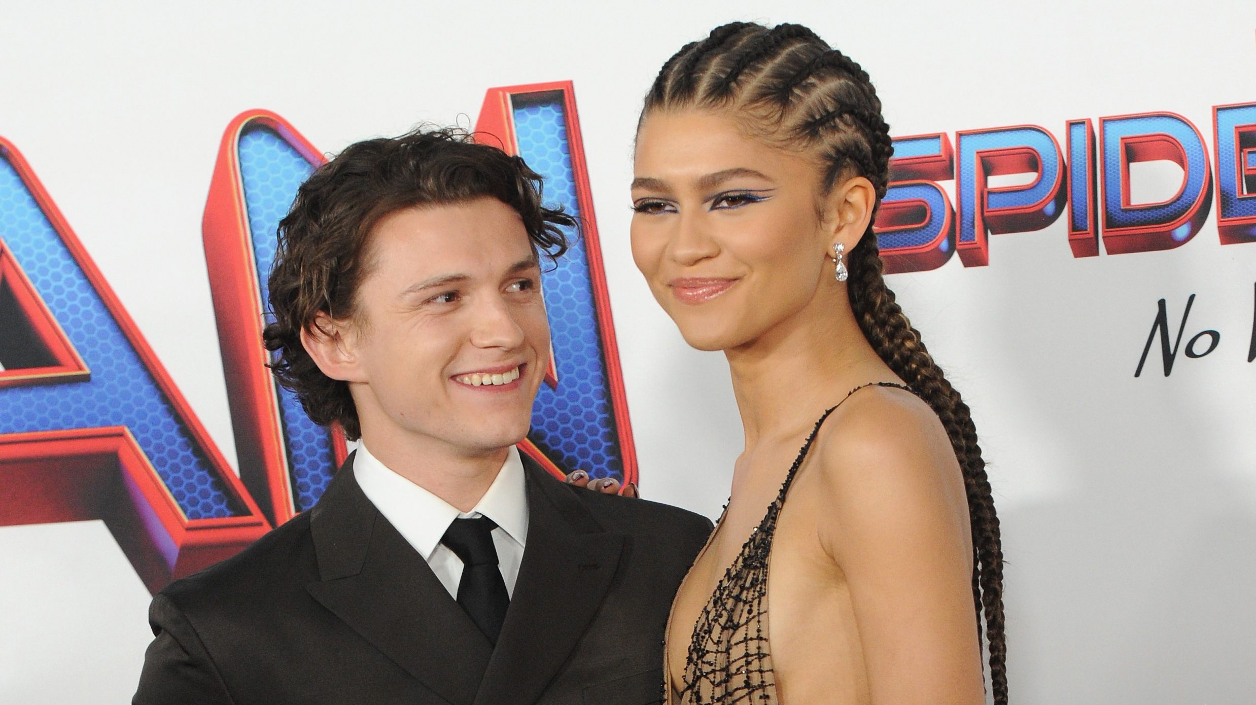 Tom and Zendaya posing for pictures.  She is wearing a nude dress with details in black and he is wearing a black suit and white shirt.