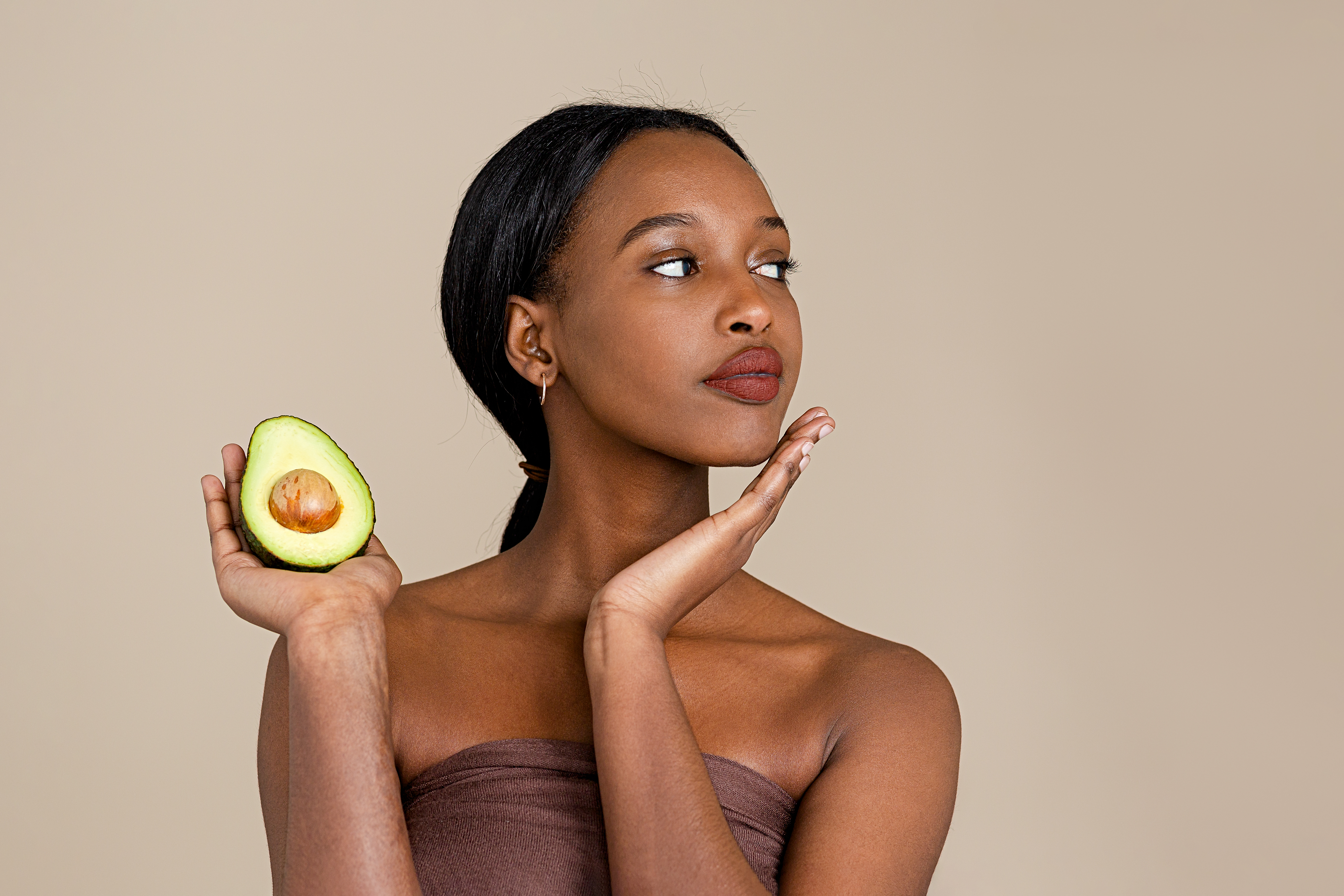 Mulher olhando para o lado com uma das mãos no queixo e a outra segurando metade de um avocado e usando toalha no corpo e com rabo de cavalo baixo