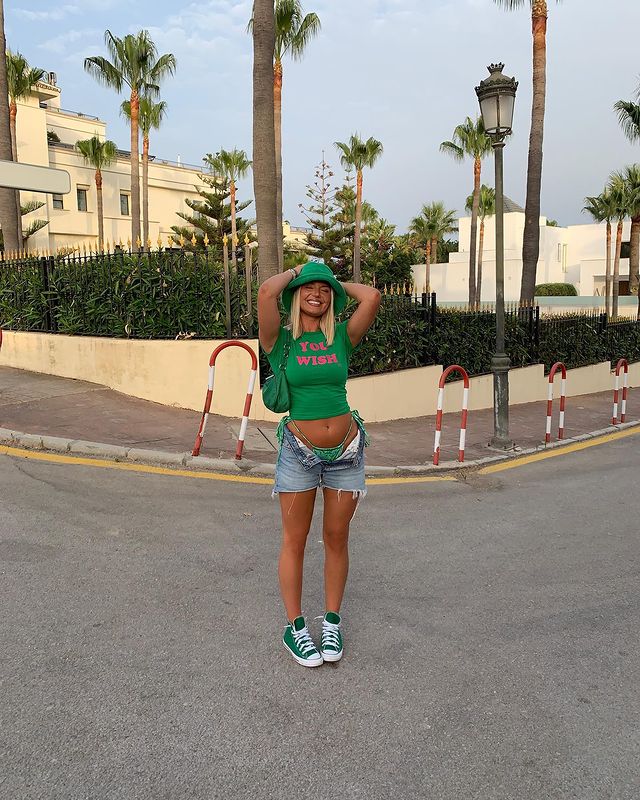 Foto de uma mulher na rua. Ela usa um chapéu de pescador verde, baby tee verde, short jeans, bolsa verde e tênis verde. Ela está com as duas mãos na cabeça, de olho fechado e sorri para a foto.