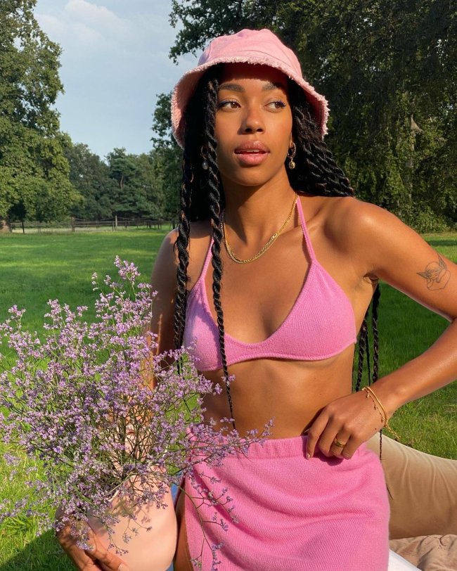 Foto de uma mulher em um gramado. Ela usa um chapéu de pescador rosa e biquíni rosa com saia rosa. Ela segura flores com a mão direita, está com a mão esquerda na cintura, olha para o lado e não sorri para a foto.