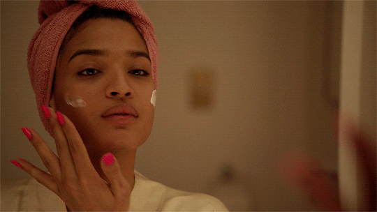 Gif da atriz Indya Moore com toalha rosa na cabeça passando creme no rosto em frente a um espelho