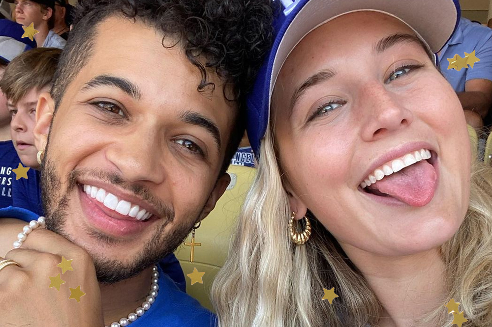 Selfie de Jordan e Ellie Fisher; ele está sorrindo com a mão segurando o queixo e ela está sorrindo e mostrando a língua; os dois estão olhando para câmera com os rostos próximos usando camiseta azul com detalhes brancos e Ellie também usa um boné das mesmas cores; eles estão sentados em uma arquibancada de cadeiras amarelas com outros torcedores ao fundo; estrelas amarelas decoram a imagem