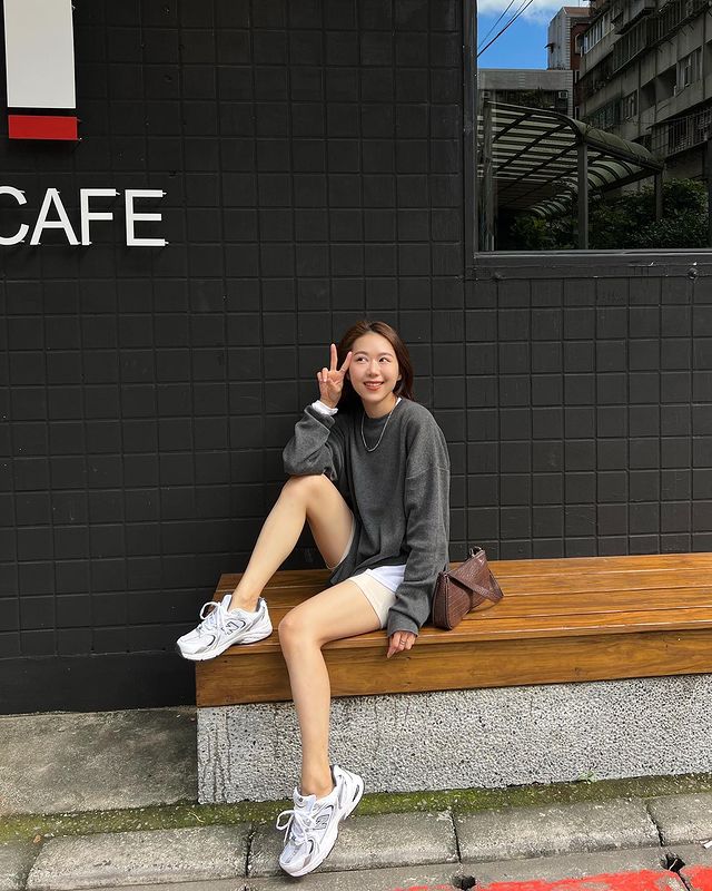 Foto de uma mulher sentada em um banco em frente a uma cafeteria. Ela usa um moletom cinza oversized, short branco, bolsa baguete marrom e tênis esportivo branco. Ela apoia a perna direita no banco, faz sinal de 