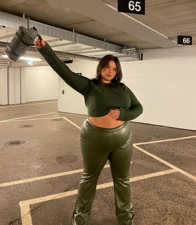 Foto de uma mulher em um estacionamento. Ela usa um top cropped de manga longa verde, calça de couro verde no mesmo tom e uma bolsa cinza. Ela está com a mão esquerda na barriga, mão direita levantada, olha para a câmera e não sorri.