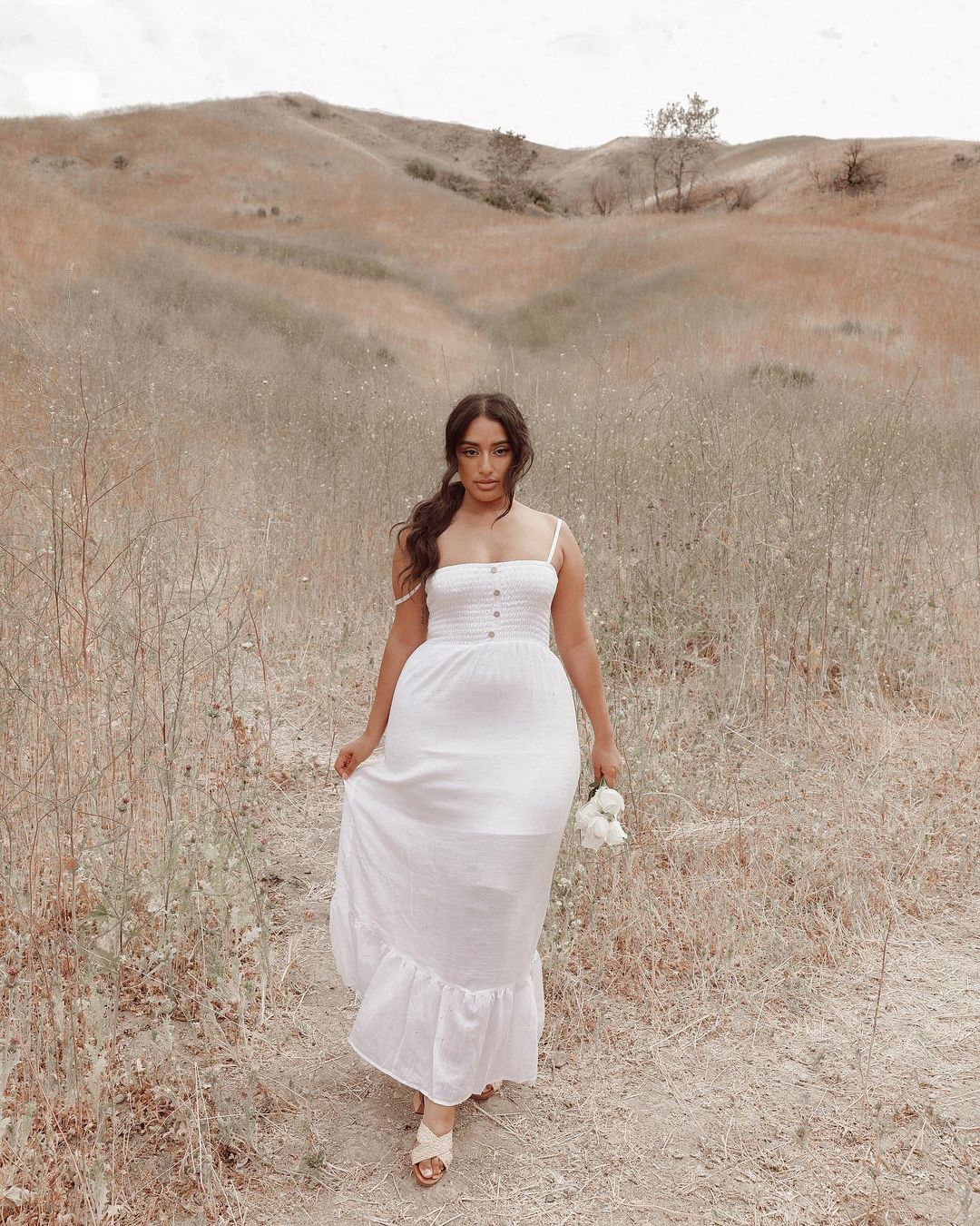 Garota usando vestido branco longo de alcinha com cabelo preso em rabo baixo em paisagem de campo