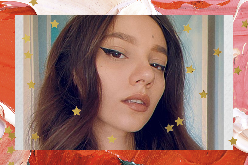 Selfie de Lorenza Vallotto com delinado preto e expressão séria olhando para câmera; a margem é uma textura de tintas em tons de vermelho, rosa, branco, laranja e verde; estrelas amarelas decoram a imagem