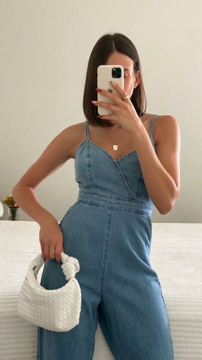 Selfie de uma mulher na frente do espelho. Ela usa um macacão jeans e bolsa branca.