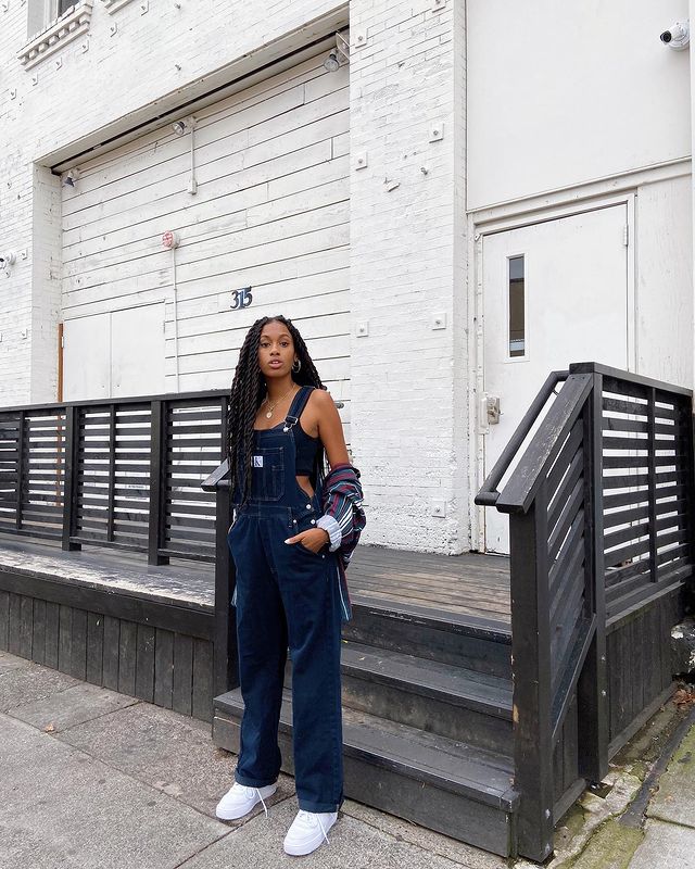Foto de uma mulher em uma calçada em frente a uma escada. Ela usa um macacão jeans, top cropped preto e segura uma camisa azul listrada. Ela está com as mãos nos bolsos do macacão, olha para a câmera e não sorri.
