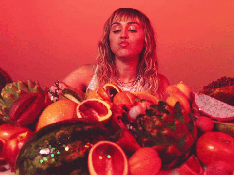 Miley Cyrus em frente a uma pilha de frutas comendo fruta