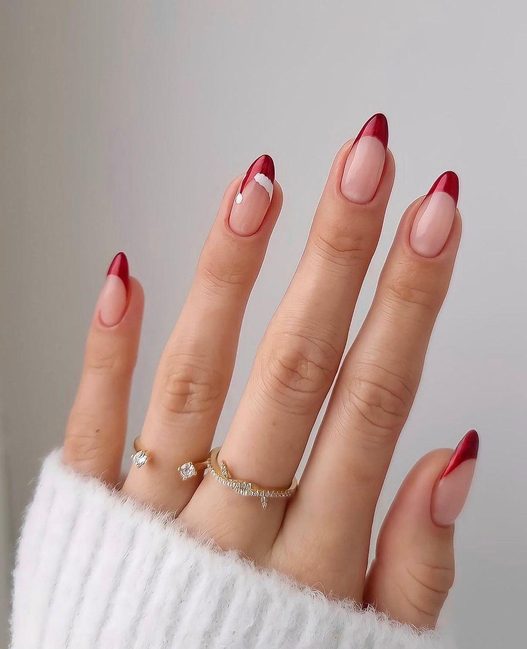Mão com unhas com francesinha vermelha de Natal, anéis dourados em dois dedos e parte de uma blusa branca de manga comprida