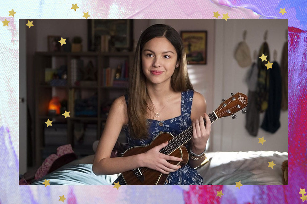 Olivia Rodrigo sentada em uma cama num quarto com prateleiras ao fundo, vestido estampado azul escuro, cabelo solto e ukulele na mão.