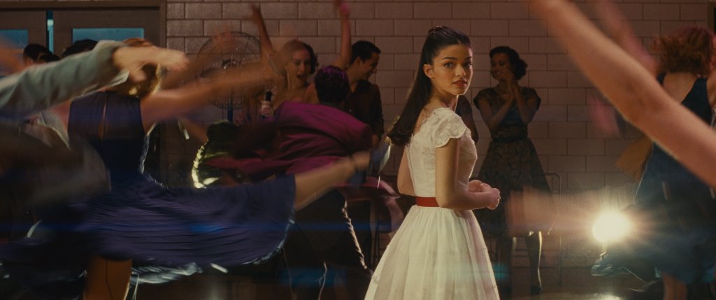 Rachel Zegler como María em Amor, Sublime Amor; ela está parada em uma pista de dança olhando para trás por cima do ombro cercada por outras pessoas usando um vestido branco com cinto vermelho com expressão surpresa