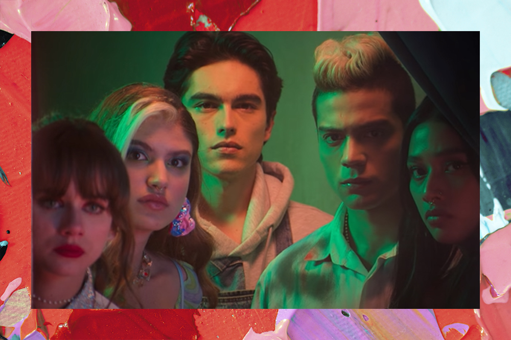 Personagens da série Rebelde em cena do trailer oficial da nova produção da Netflix; eles estão reunidos com expressões tristes e confusas observando algo parados atrás de uma cortina iluminados por uma luz verde; a margem é uma textura de tintas nas cores rosa, vermelho, azul, verde, roxo e branco