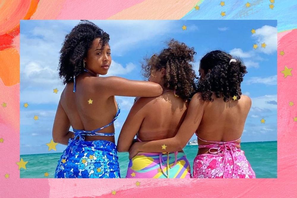 Montagem com o fundo rosa e azul com detalhe de estrelinhas douradas com a foto de três mulheres. As três mulheres estão de costas para a foto na praia e usam um conjunto de biquíni com saia com estampa combinando. Apenas a mulher da esquerda olha para a foto e não sorri.
