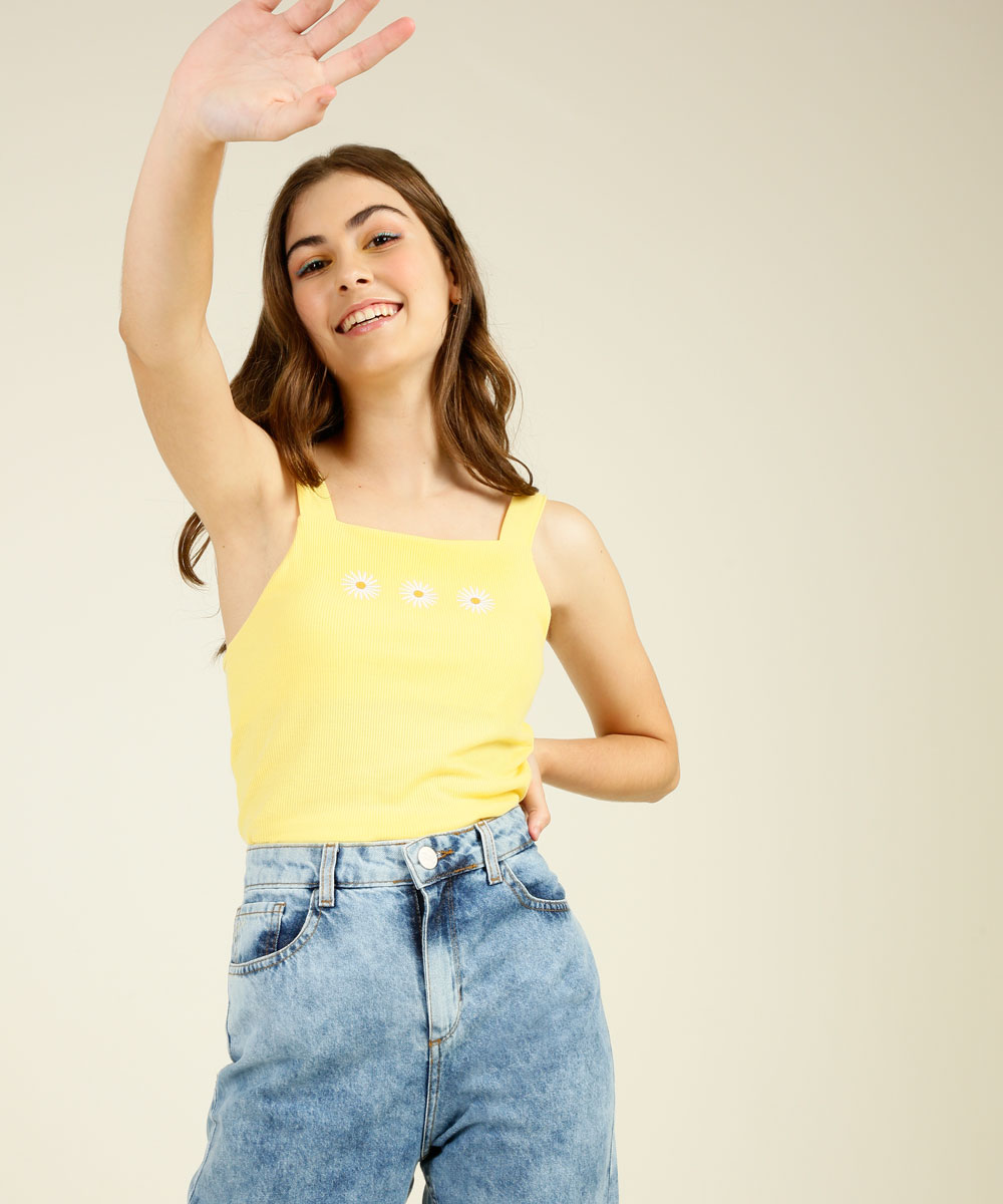 Garota usando top cropped amarelo com flores e calça jeans, sorrindo, com uma das mãos na cintura e a outra esticada para cima