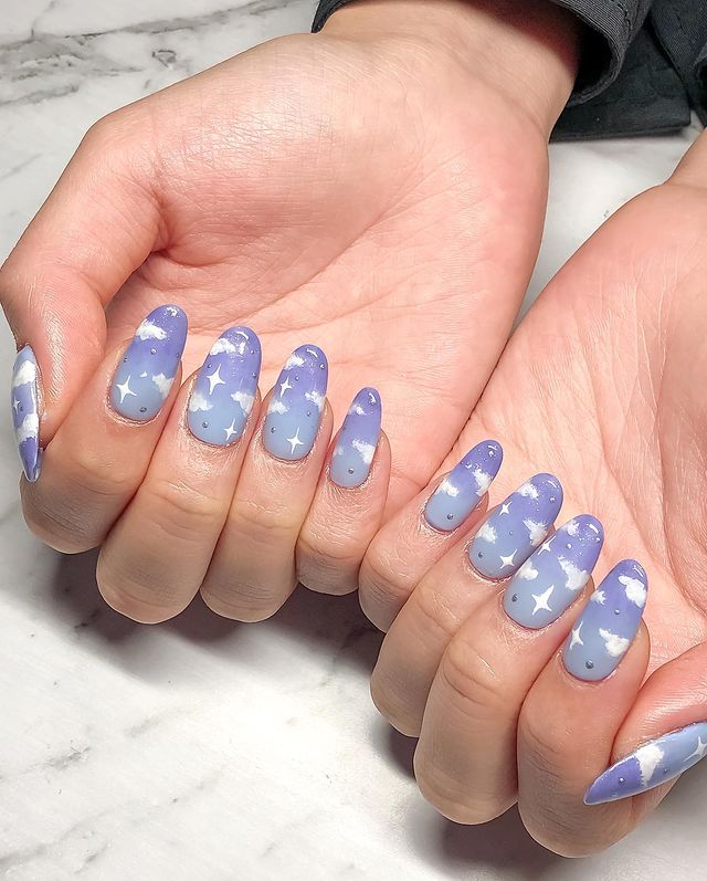 Foto de mãos com as unhas decoradas com esmalte lilás e desenho de nuvens brancas.