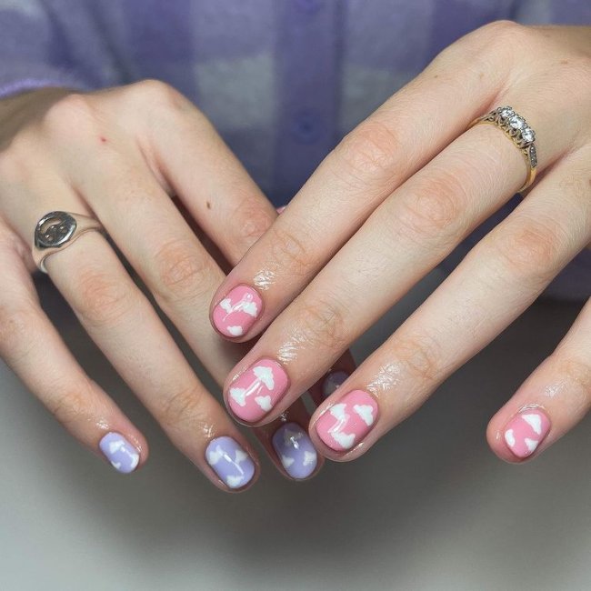 Foto de mãos com esmalte lilás e rosa com desenho de nuvens lilás.