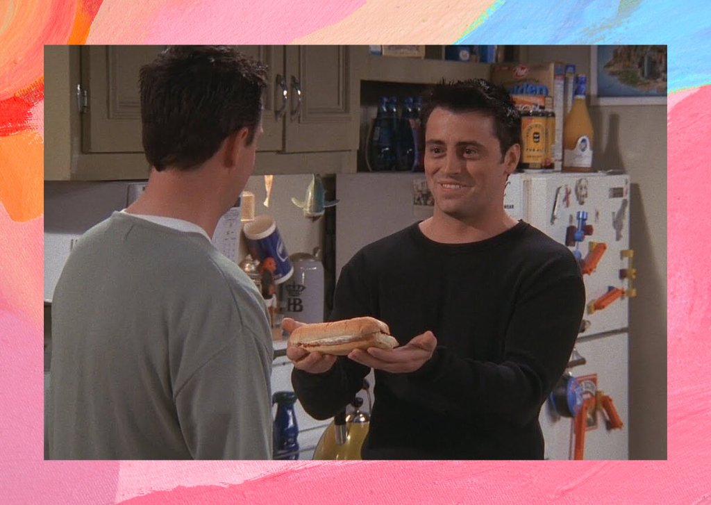 Um homem de costas e o outro com um sanduíche na mão em uma cozinha.