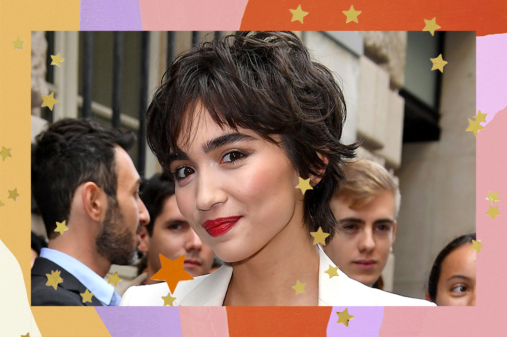 Rowan Blanchard usa blazer branco com top preto de renda, batom vermelho e o cabelo com o corte bixie cut. Ela olha para a câmera com expressão sorridente.