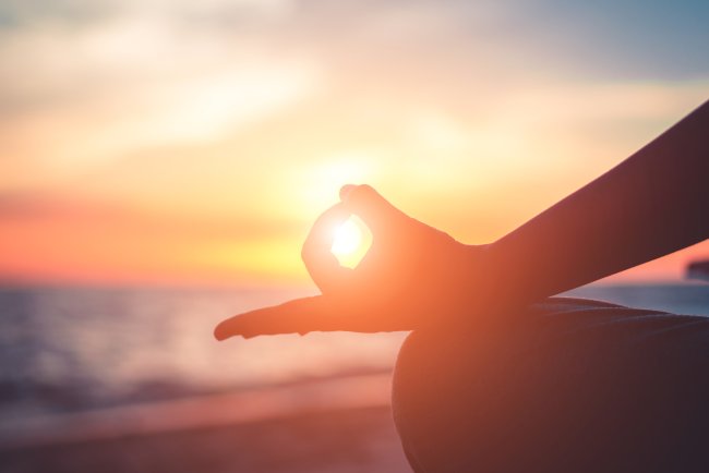 Foto de uma mão em posição de ohm, da meditação. Atrás dela, um pôr do sol