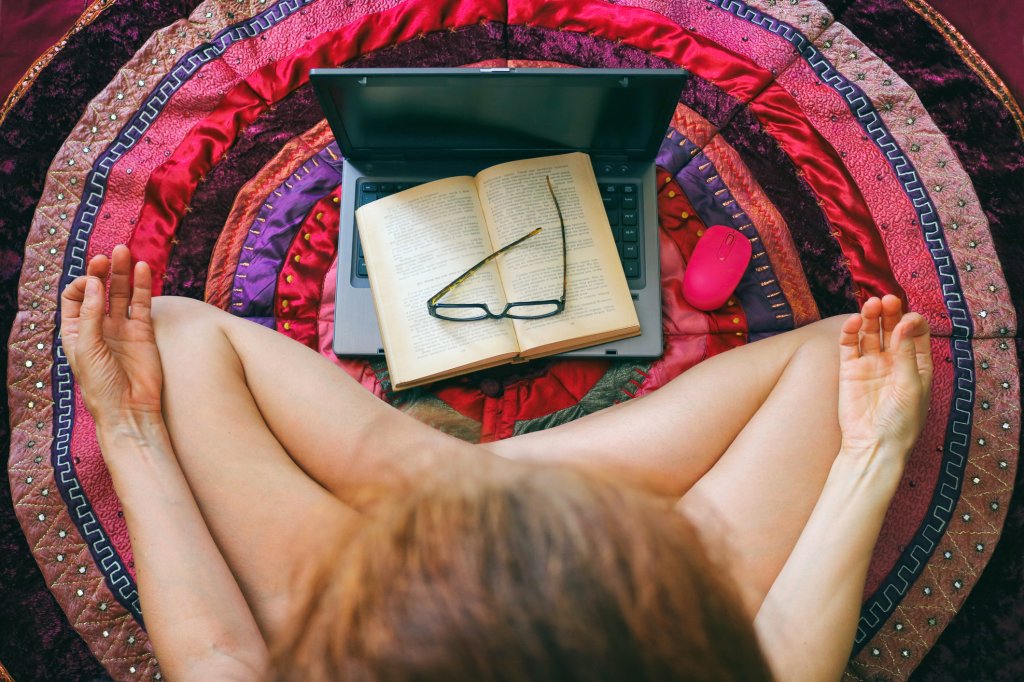Foto de uma mulher sentada no chão, com as pernas cruzadas, com os dedos em posição de meditação, em frente a um notebook. Em cima dele, tem um livro e um óculos. O tapete é bem colorido.